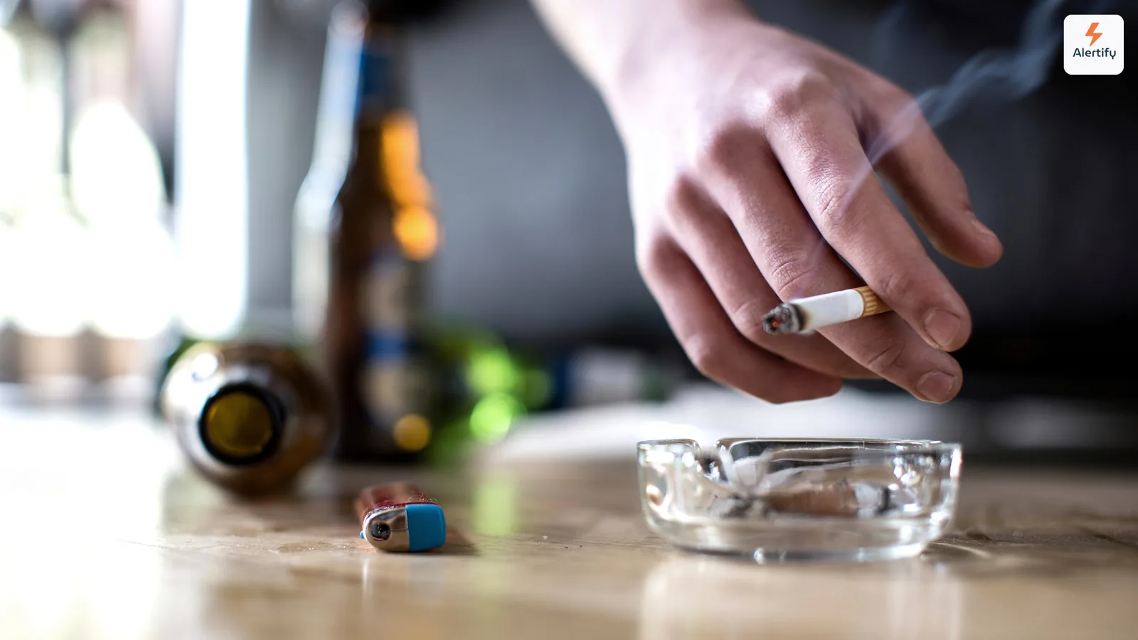Gast met een brandende sigaret boven een glazen asbak naast lege flessen op een tafel