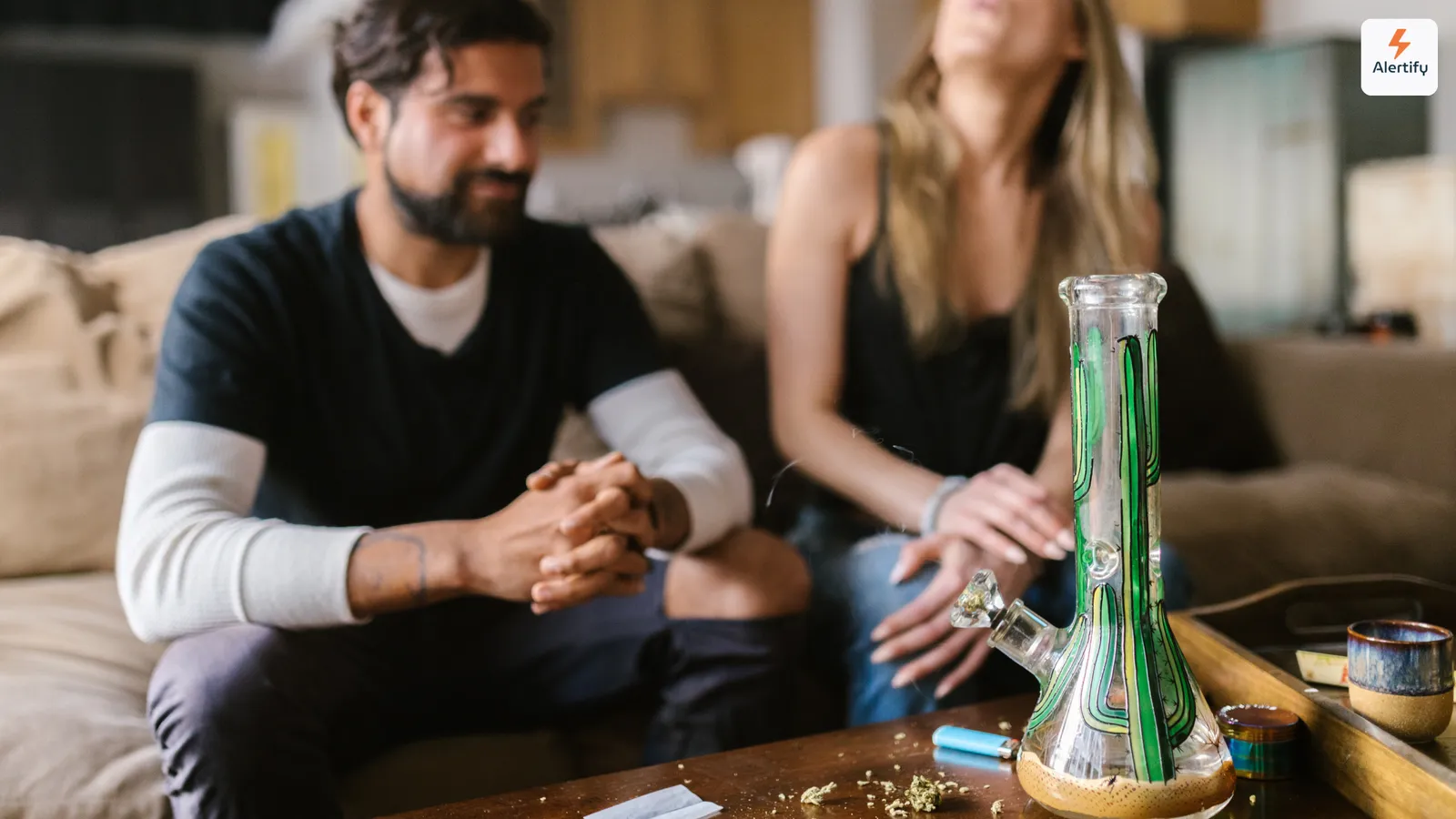 Twee gasten op een bank in een woonkamer van een vakantiewoning, met een glazen bong, een aansteker en een grinder op de salontafel voor hen.