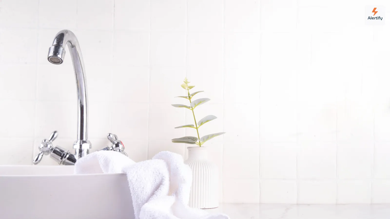 Een lichte, wit betegelde badkamer met een wasbak, een opgevouwen handdoek en een kleine plant in een pot — de soort vochtige ruimte waar schimmel het eerst toeslaat.